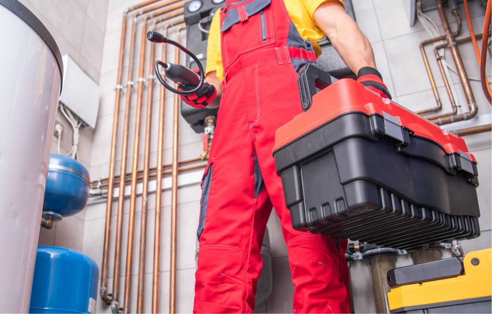 Worker in red overalls carrying a large toolbox in a utility room with pipes and tanks
