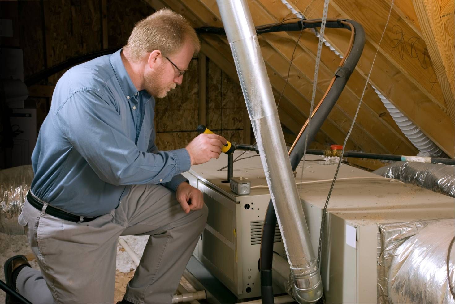 Technician inspecting HVAC equipment in an attic with ductwork and insulated piping
