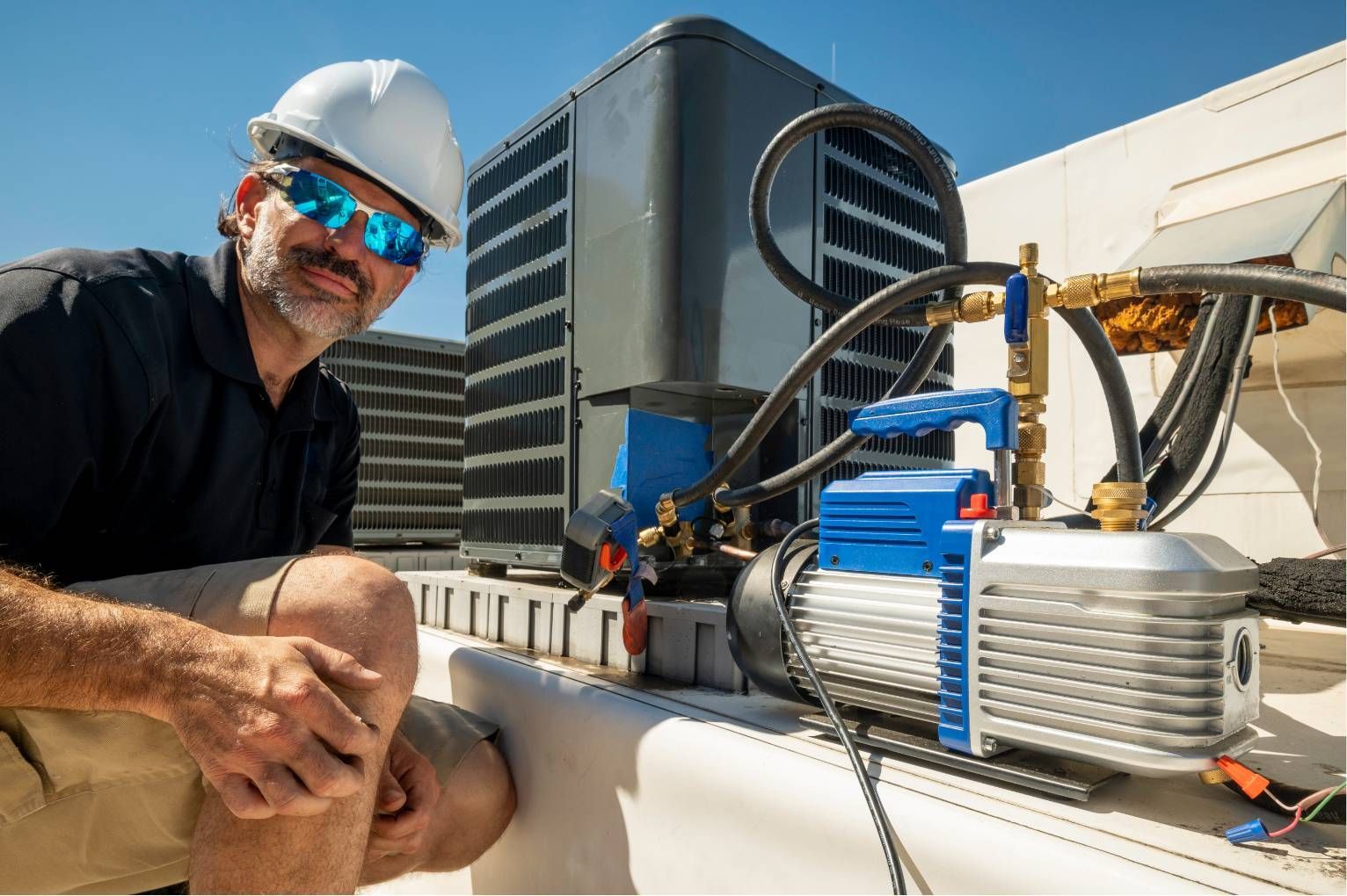 Worker on rooftop beside HVAC equipment, wearing hard hat and sunglasses, with city buildings in background