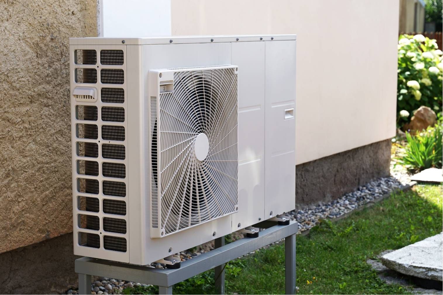 Outdoor air conditioning unit beside a building wall on a metal stand in a yard