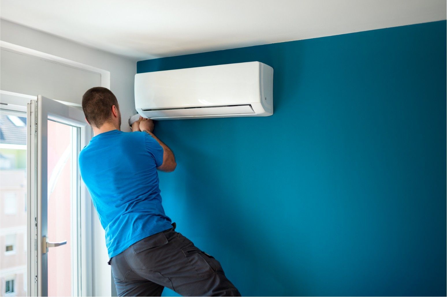 Technician installing a white wall-mounted air conditioner on a teal wall