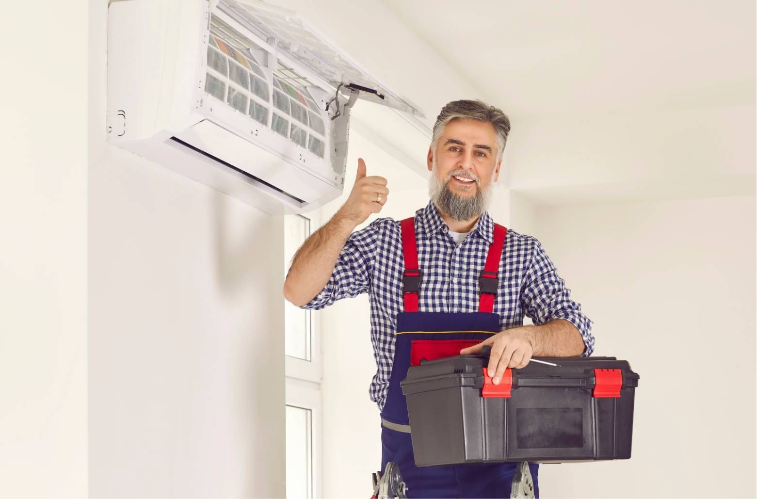 Technician in blue overalls holds a toolbox and gives a thumbs-up beside a wall-mounted air conditioner