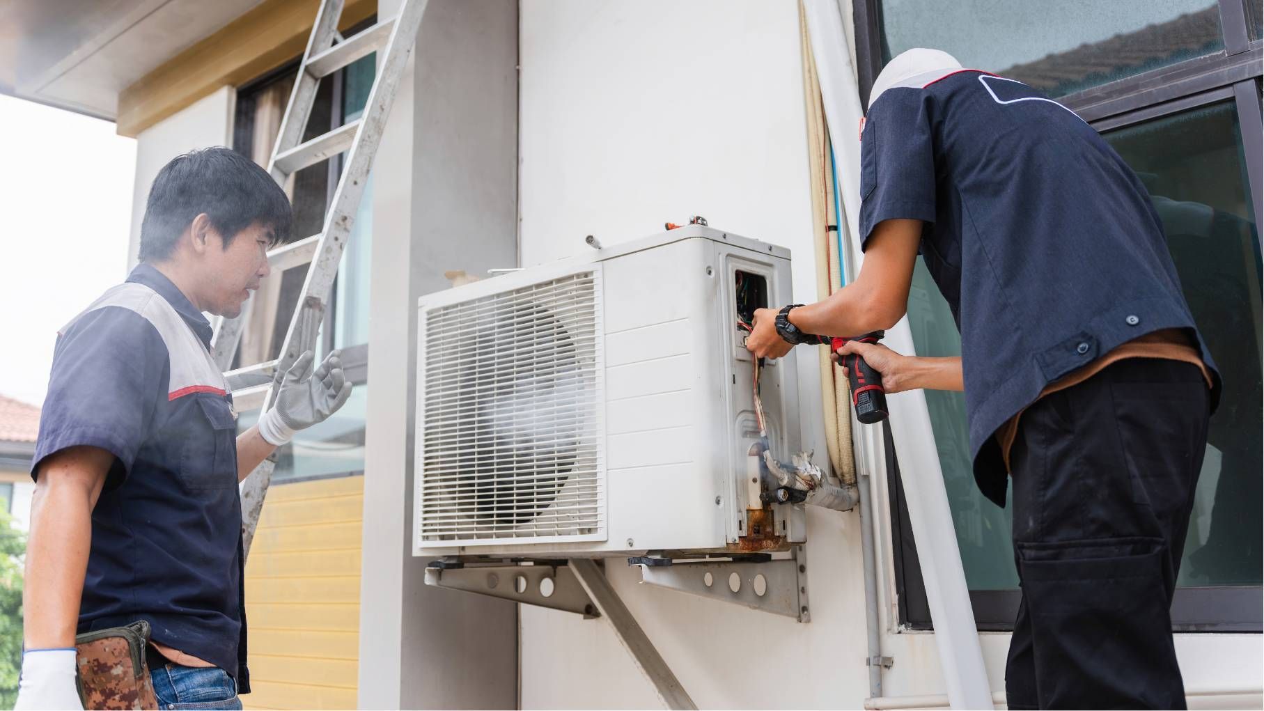 Two technicians repair an outdoor air conditioner unit mounted on a building wall.