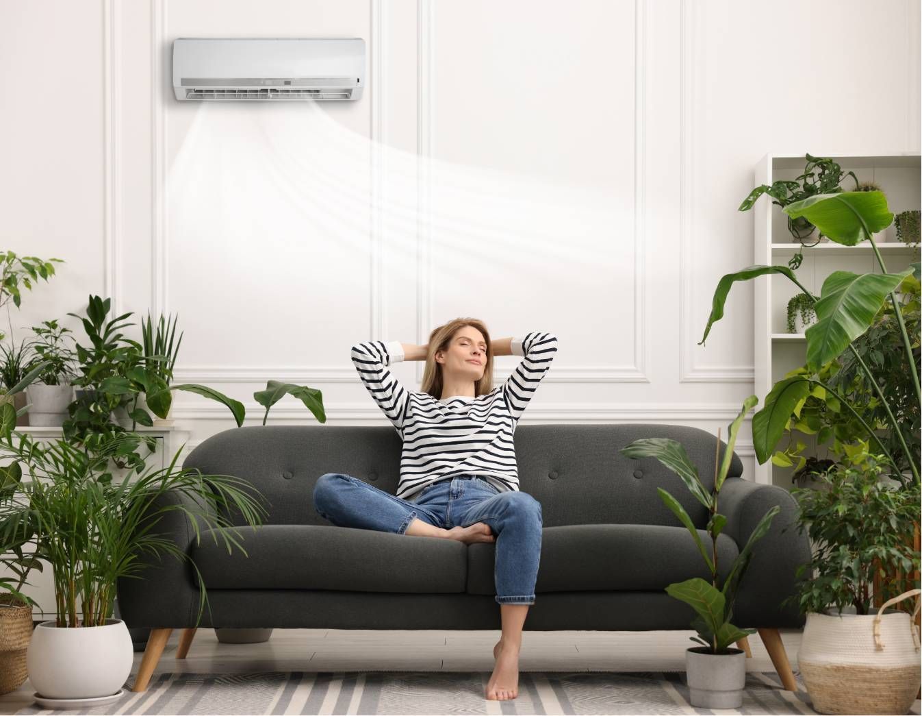 Person relaxing on a gray sofa in a bright living room with potted plants and a wall air conditioner.