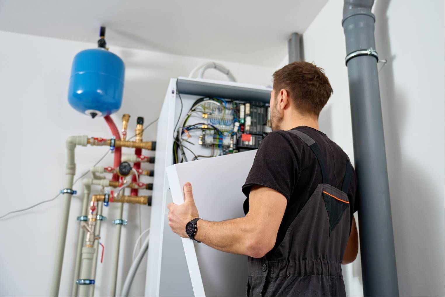 Technician inspecting an open boiler unit in a utility room with pipes and a blue tank
