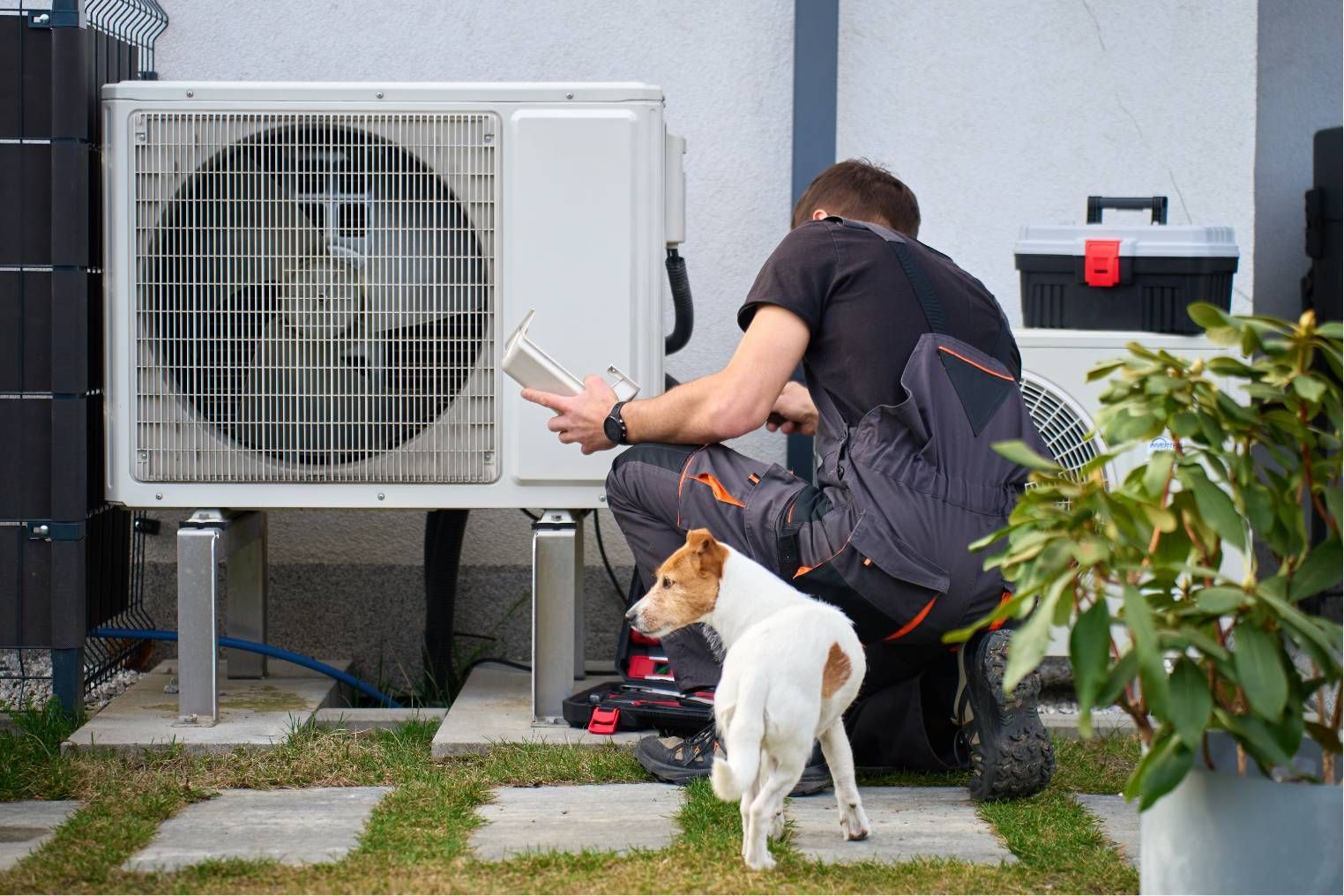 Technician kneels beside an outdoor AC unit while a small dog stands nearby in a yard.