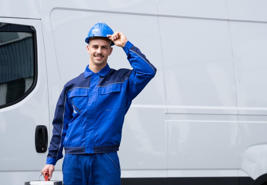 Smiling mechanic in blue uniform and hard hat standing beside a white van, tipping his helmet