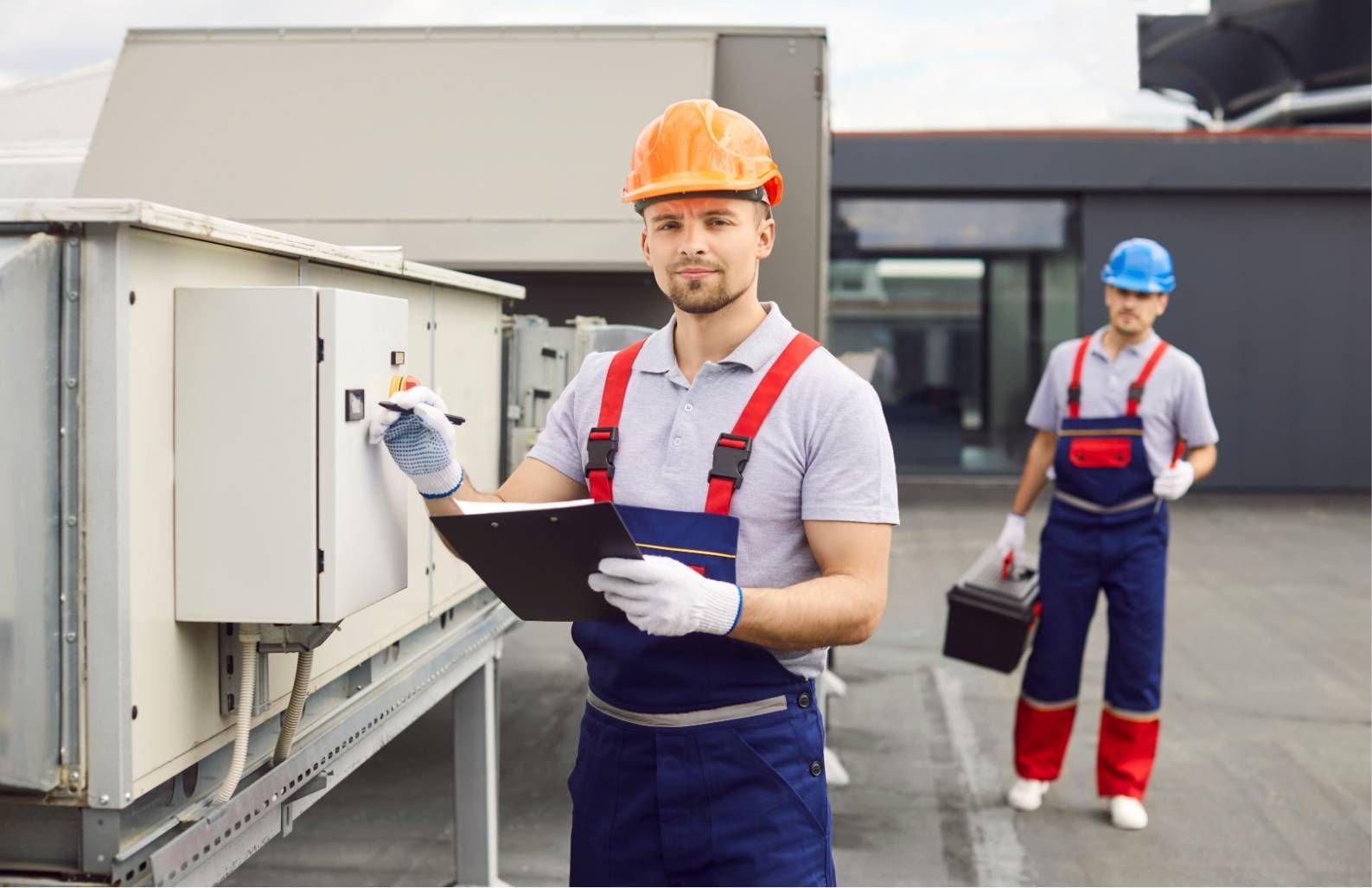 Two workers in hard hats inspect rooftop HVAC equipment, one holding a clipboard.