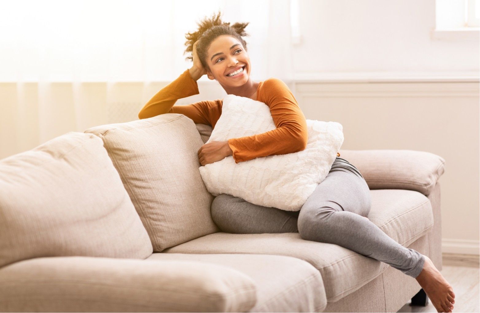 Person relaxing on a beige sofa, smiling and hugging a white pillow in a bright living room.