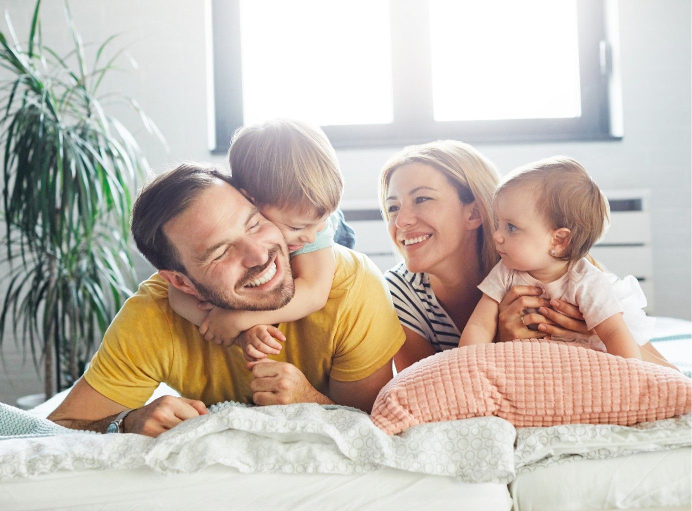 Family smiling on a bed, with two adults and two small children in a bright bedroom