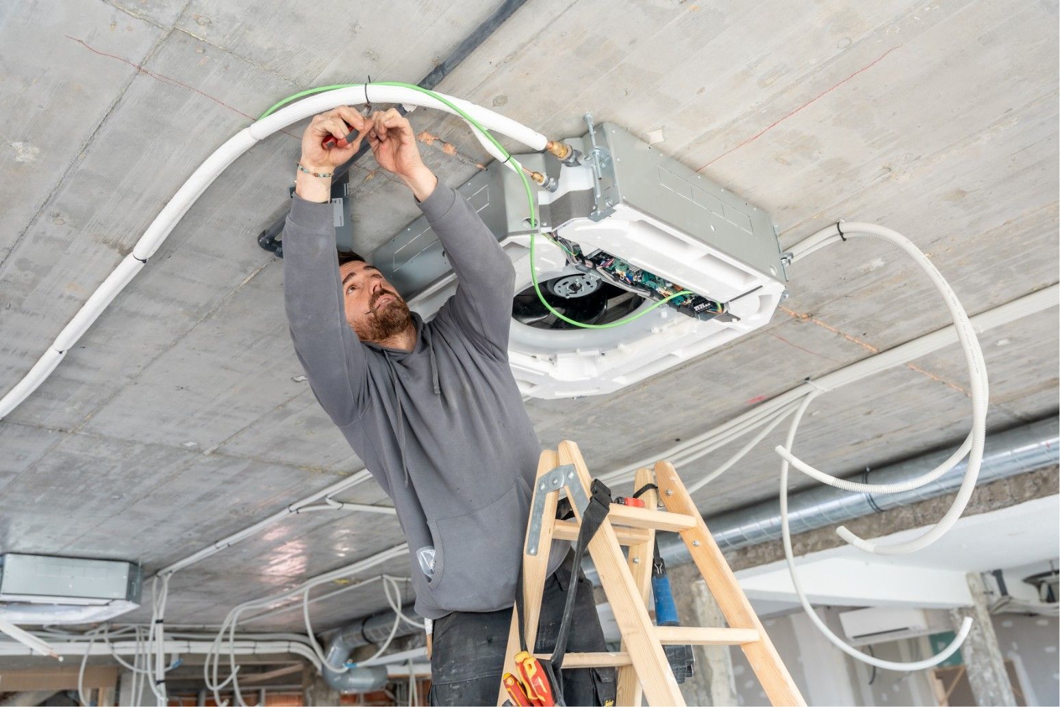 Worker wiring a ceiling-mounted HVAC unit while standing on a ladder in a concrete room.