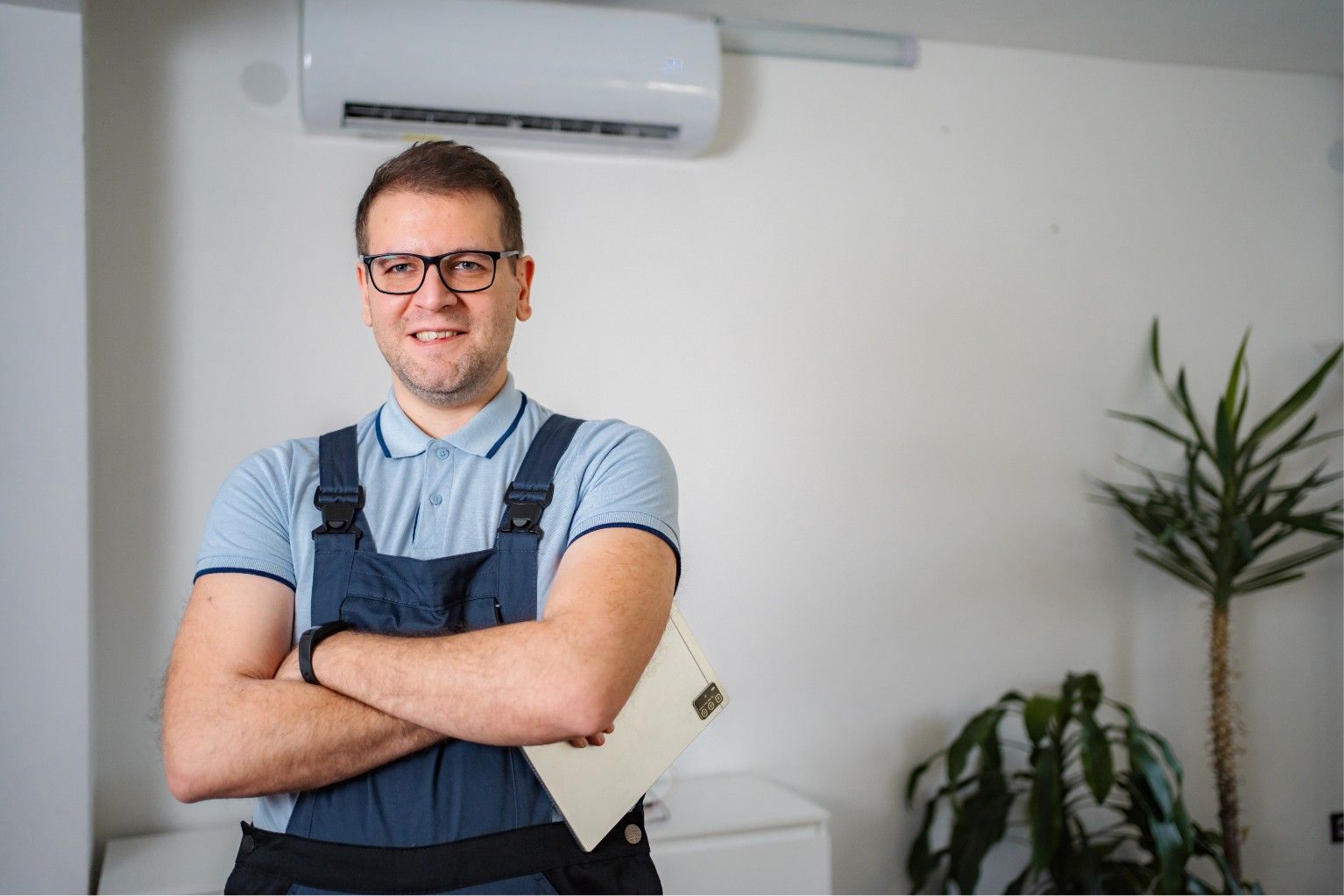 Man standing with arms crossed in an office, wearing glasses and an apron, with a plant and air conditioner behind him