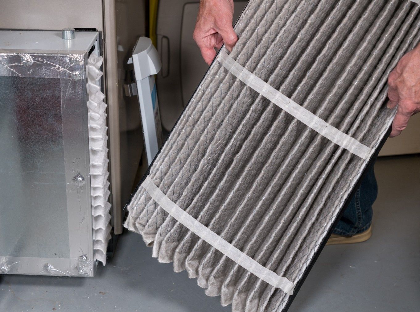 Hands holding a pleated air filter beside a metal HVAC unit indoors