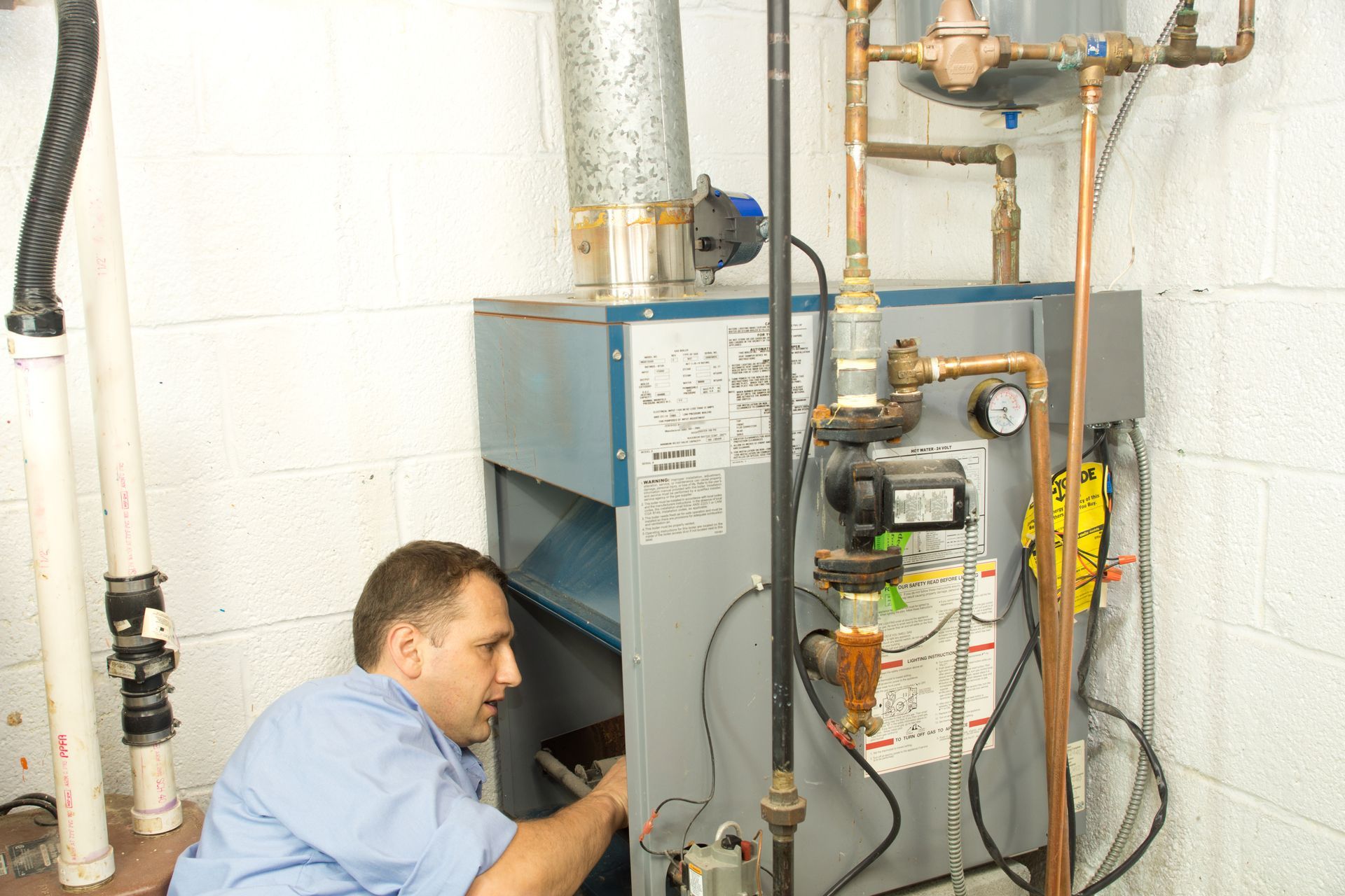 Technician inspecting a furnace and exposed pipes in a utility room, kneeling beside equipment.
