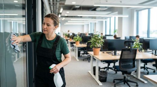 Person in overalls and green shirt cleans glass wall in bright modern office with desks computers and plants