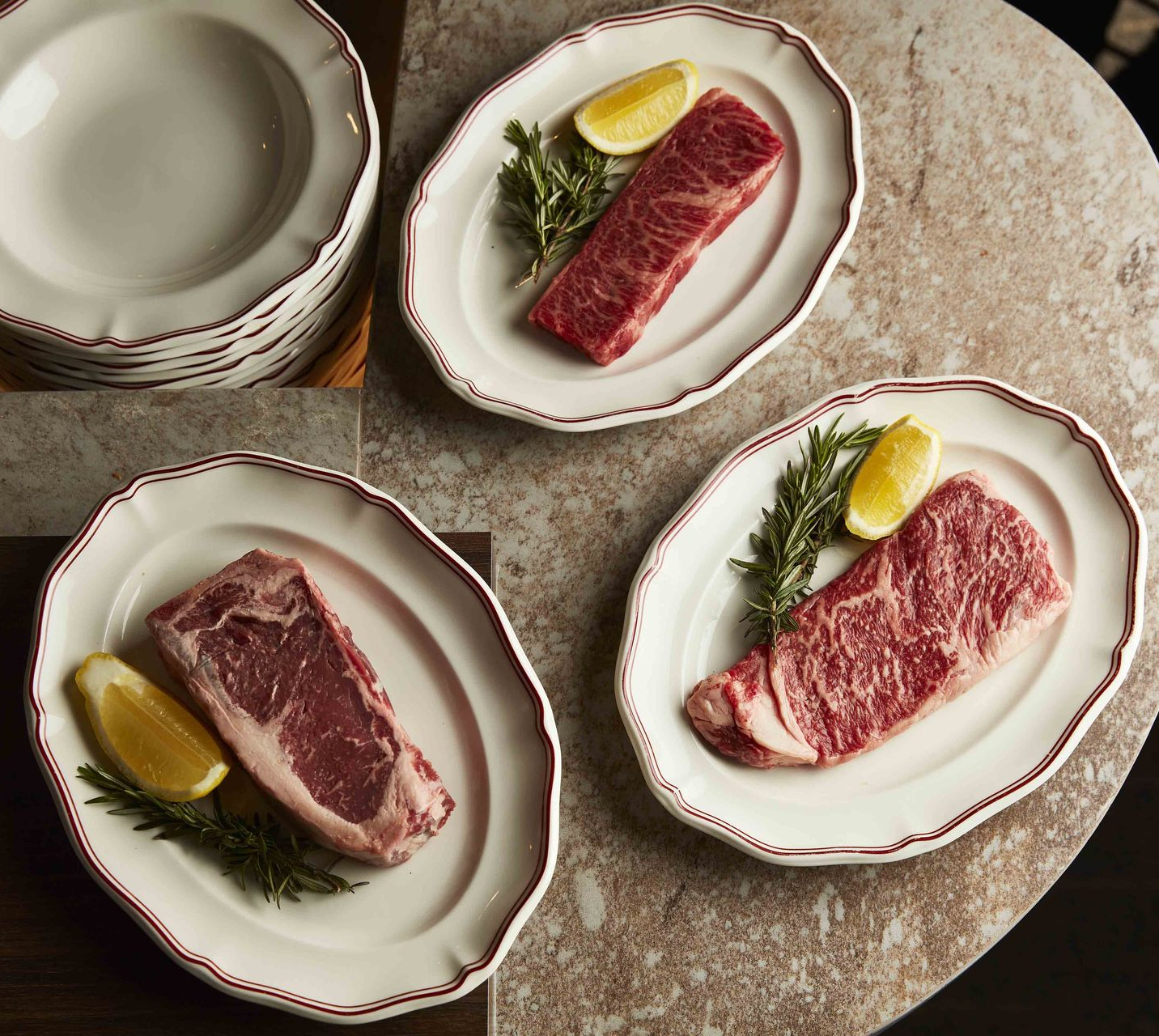 Three raw steaks arranged vertically on white plates against a dark tabletop.