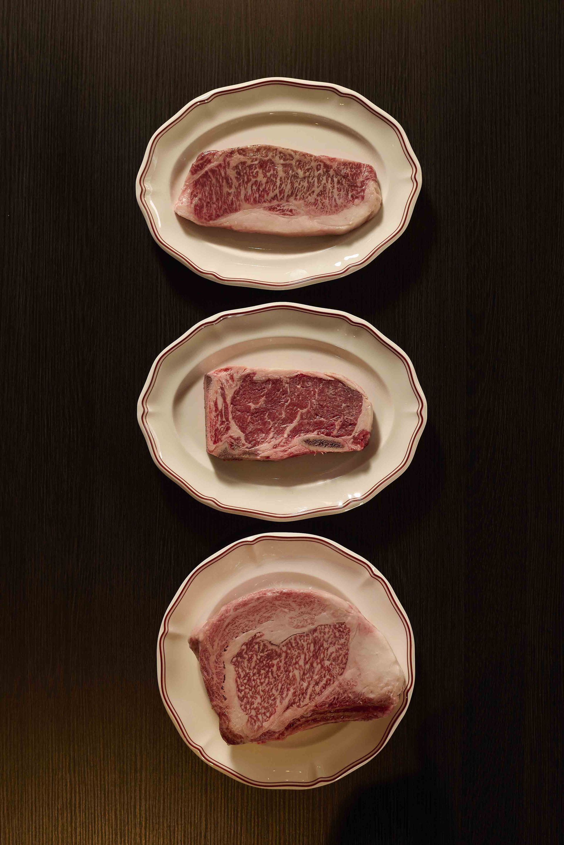 Three raw steaks arranged vertically on white plates against a dark tabletop.