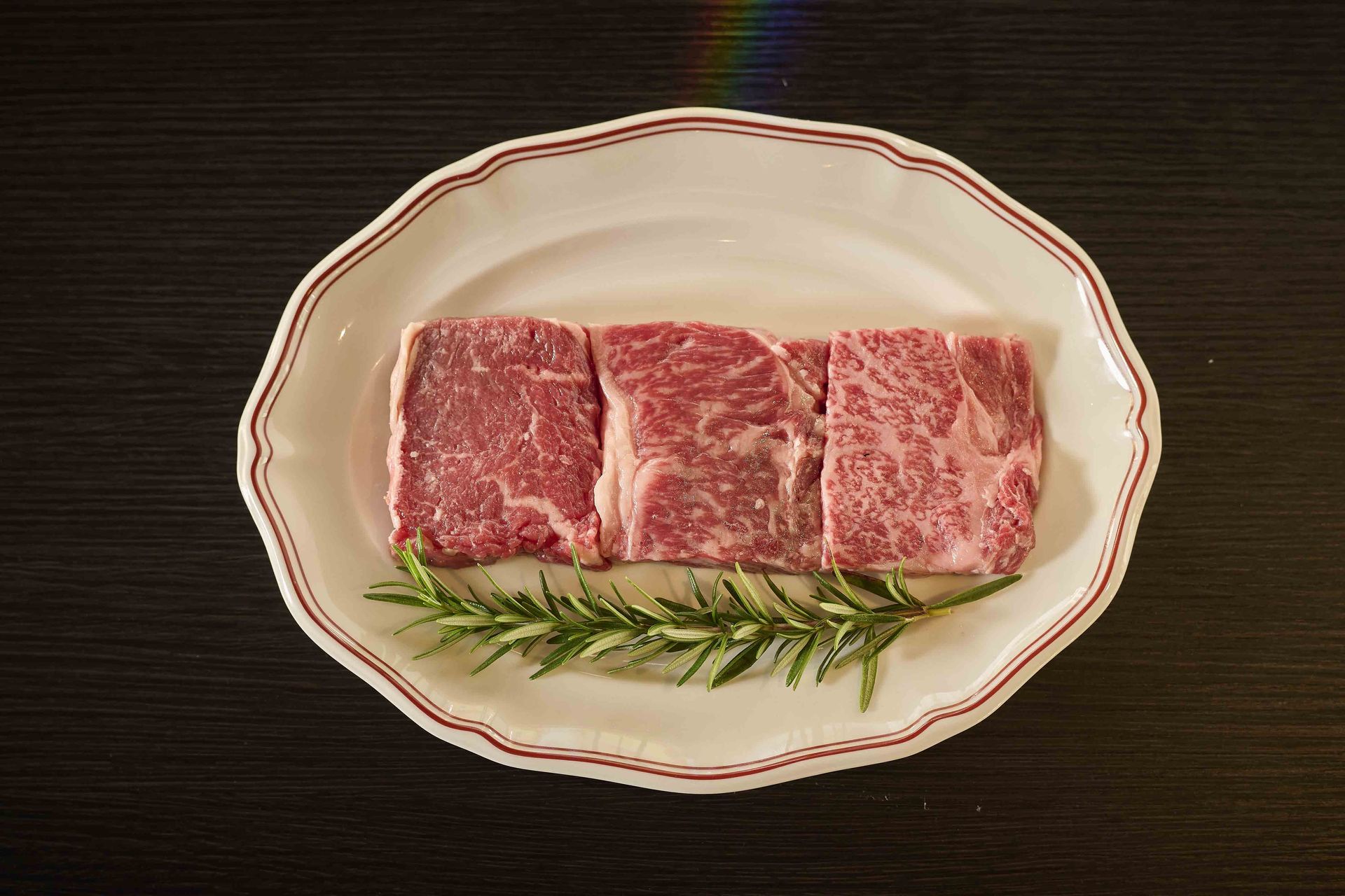 Raw steak on a white plate with rosemary sprigs on a dark table