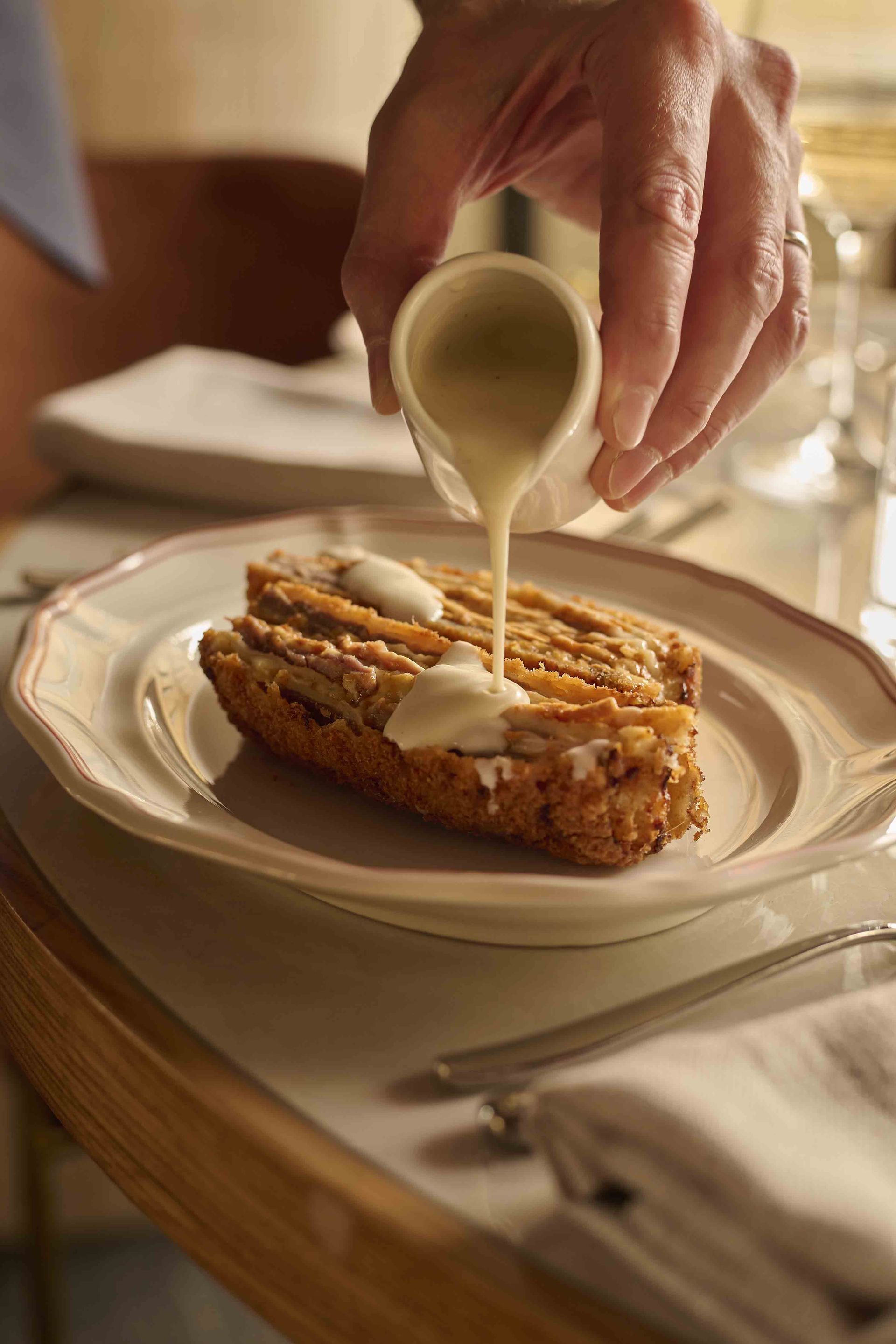 Hand pouring cream over a slice of cake on a white plate at a table