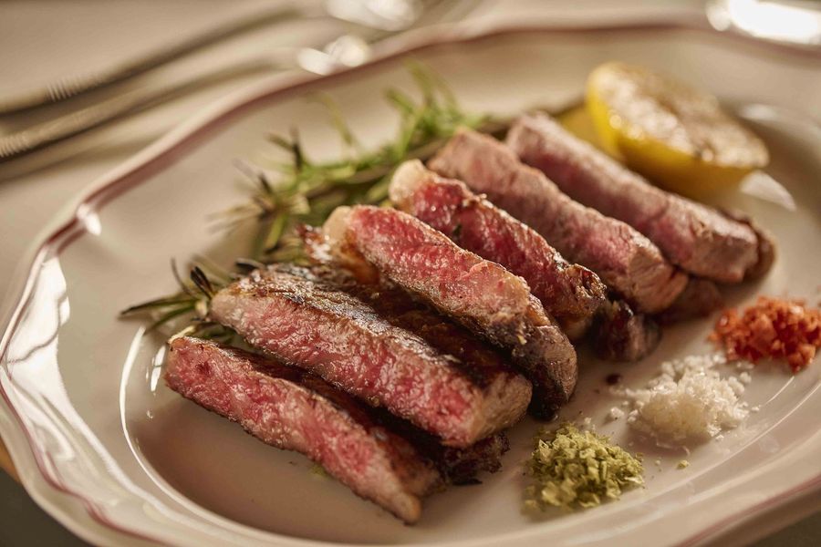 Sliced seared steak on a white plate with herbs, sauces, and a lemon wedge