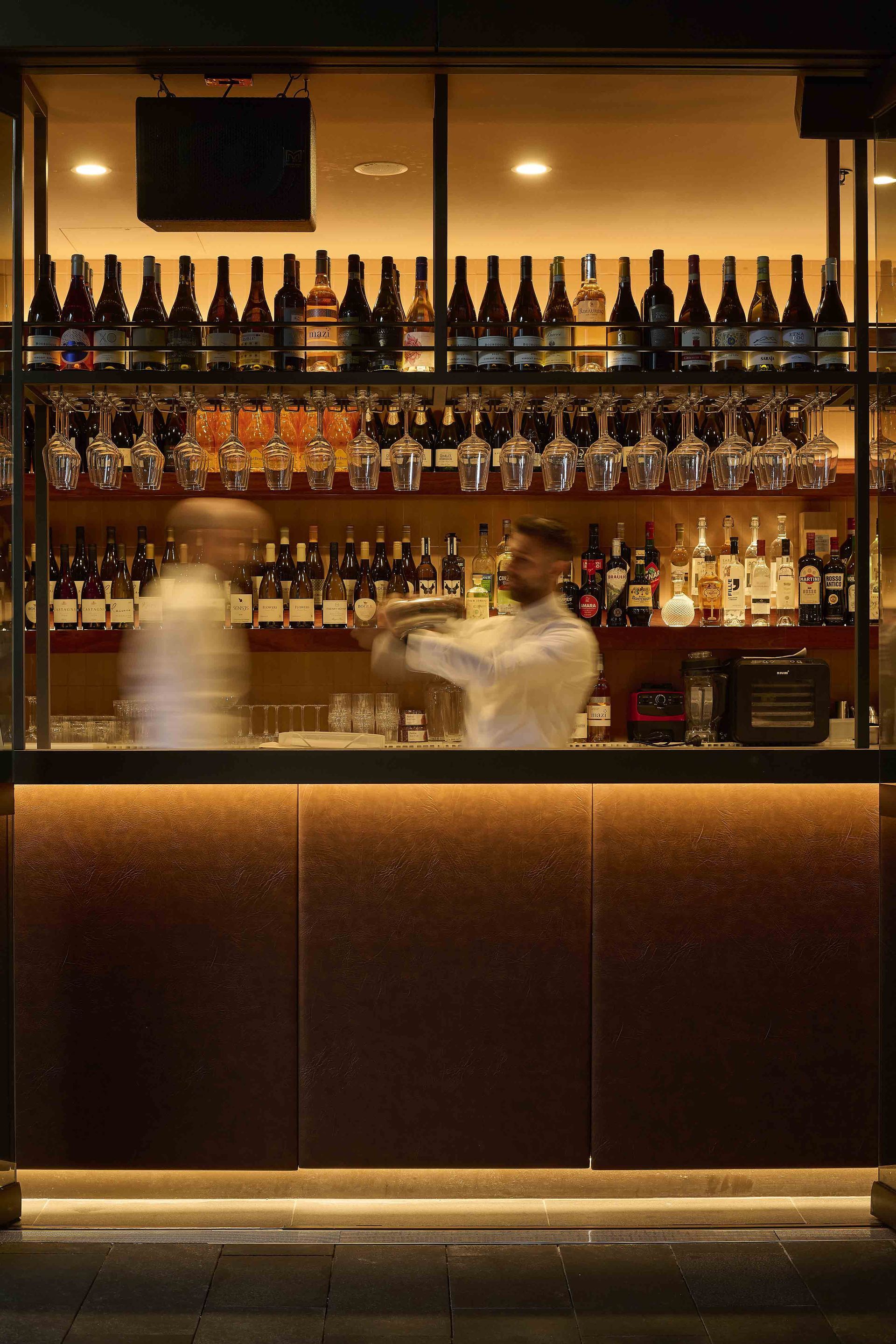 Warmly lit bar interior with shelves of bottles and two blurred bartenders behind the counter