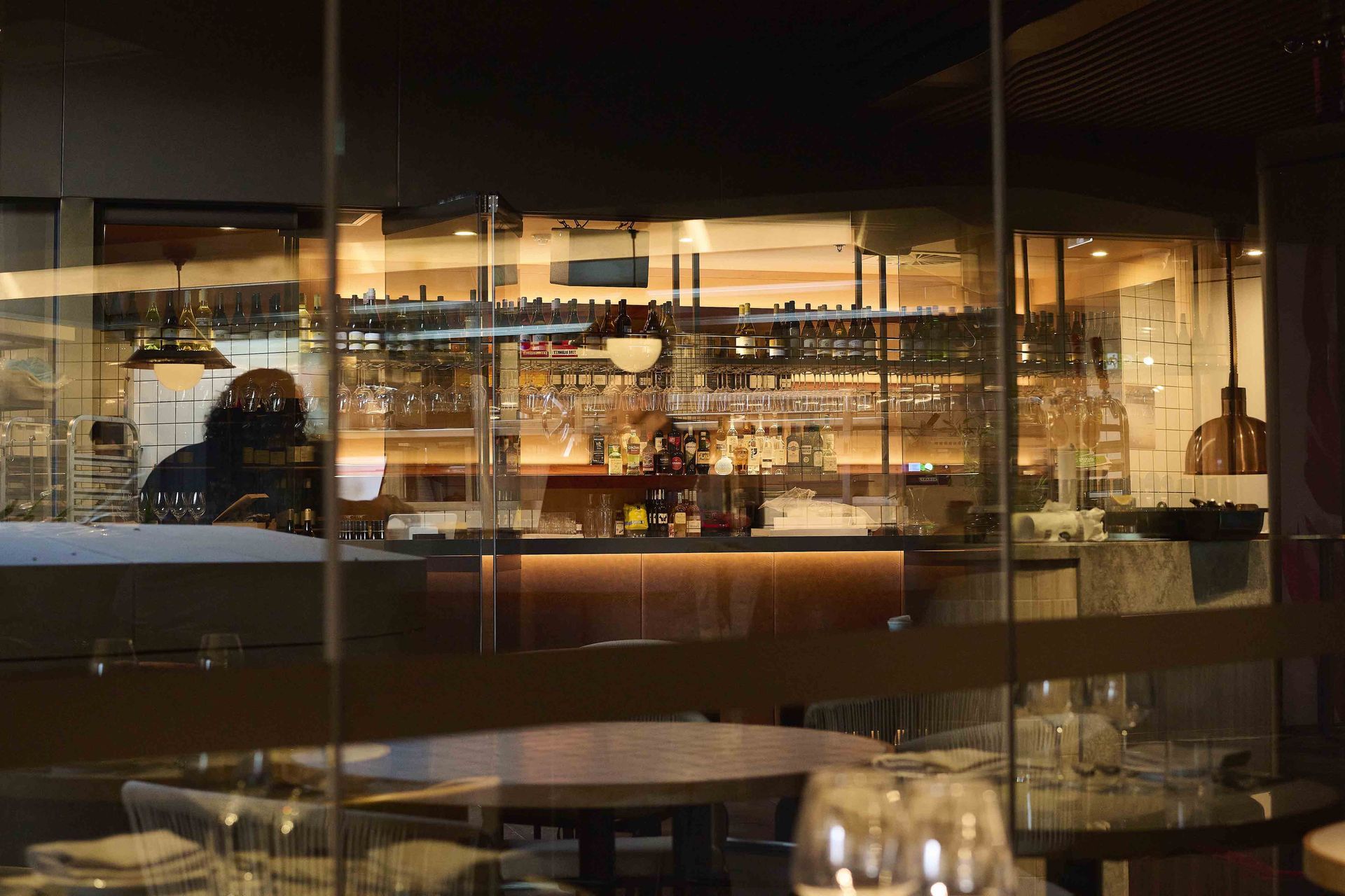 Dimly lit restaurant interior with a reflective glass wall, warmly lit bar, and empty tables in the foreground