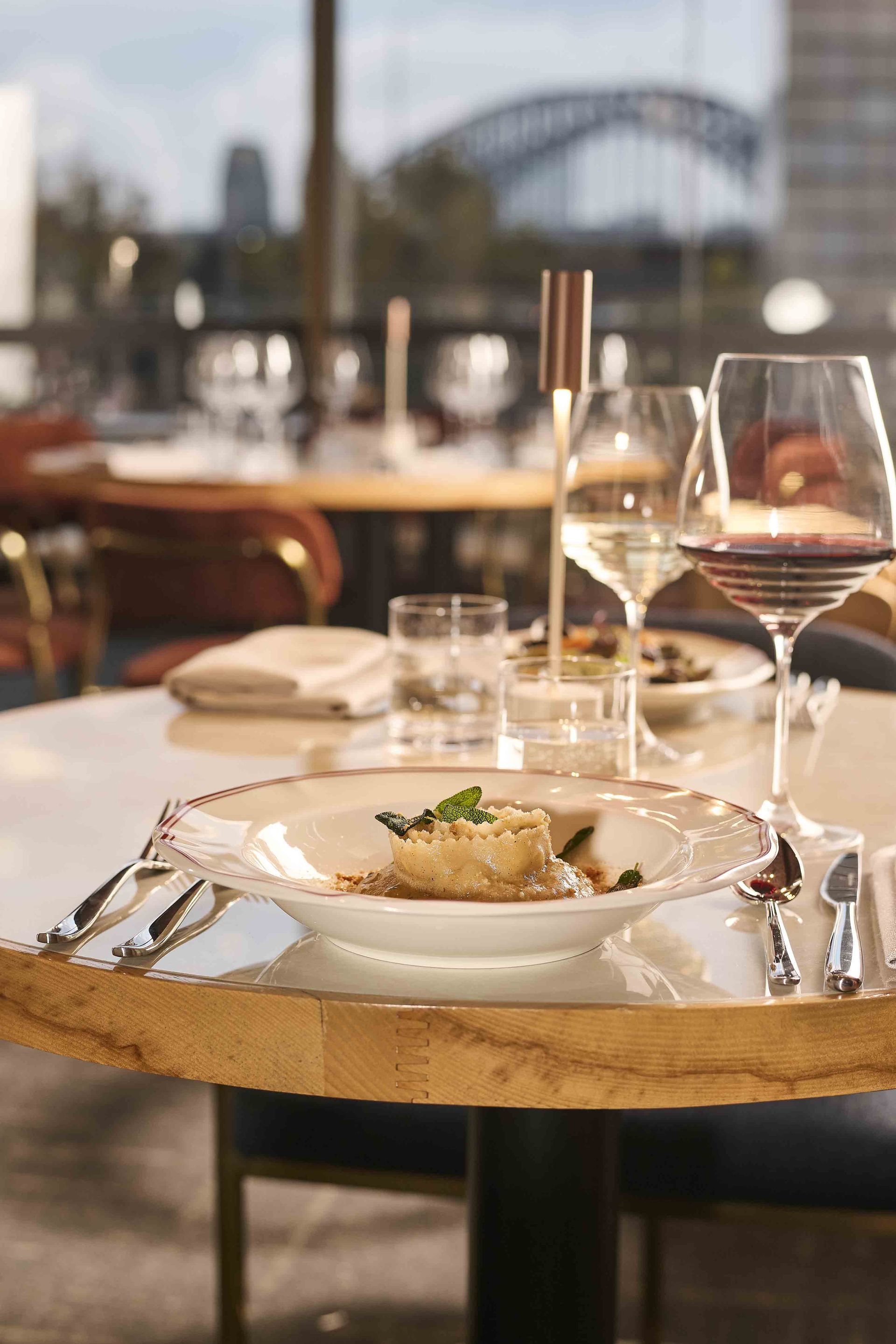 Restaurant table set for a meal, with wine glasses and a plated dish in front of a city view.