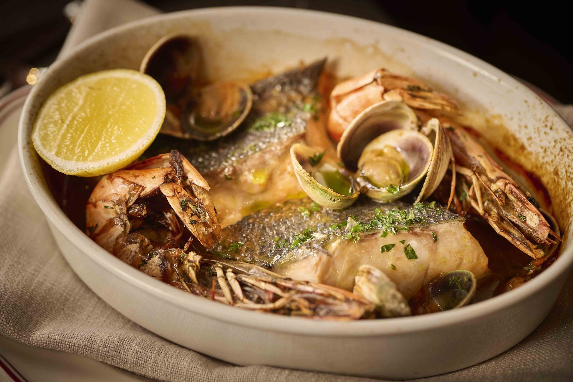 Seafood stew in a white bowl with clams, fish, and a lemon wedge