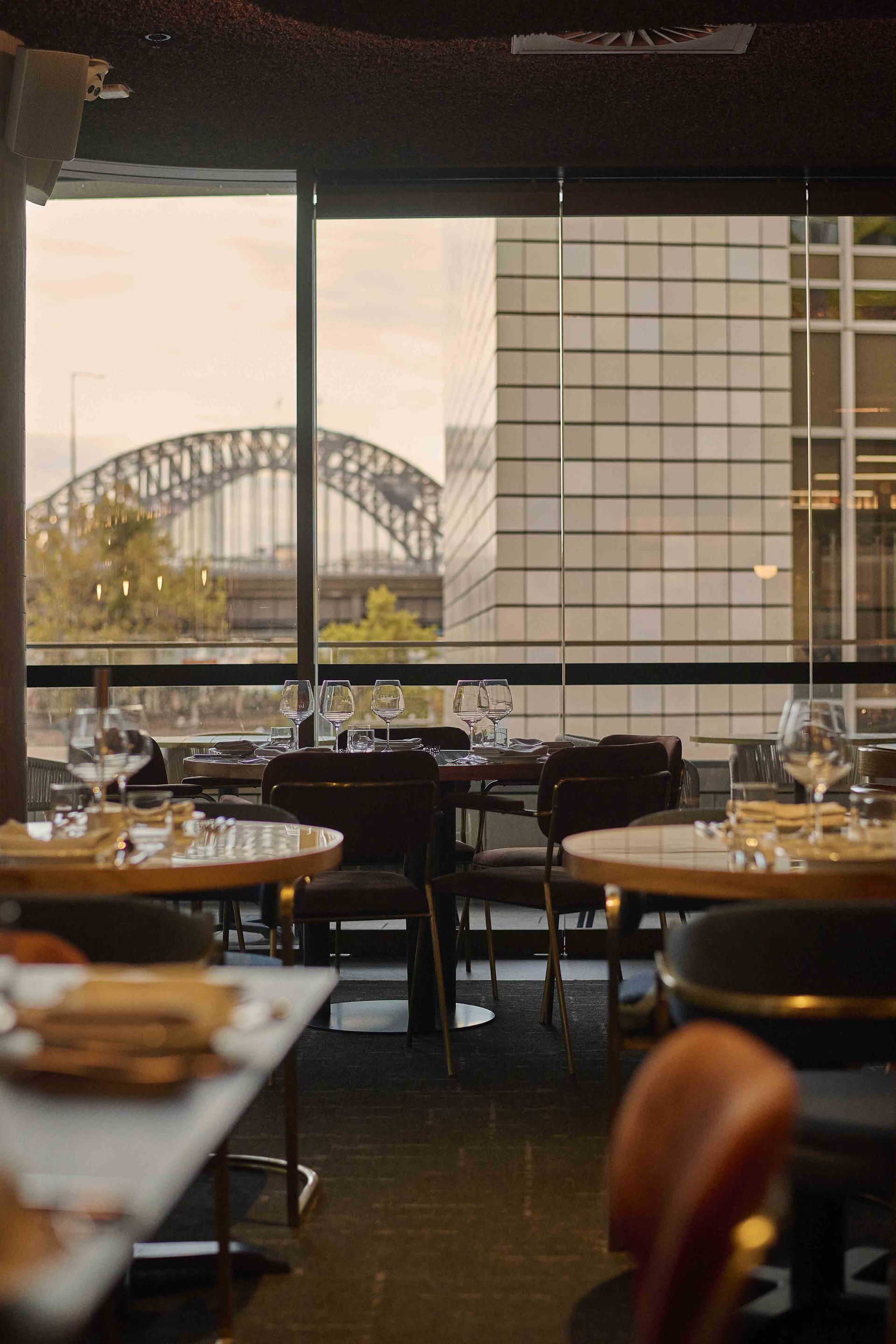 Empty restaurant with tables and chairs overlooking a city bridge through large windows