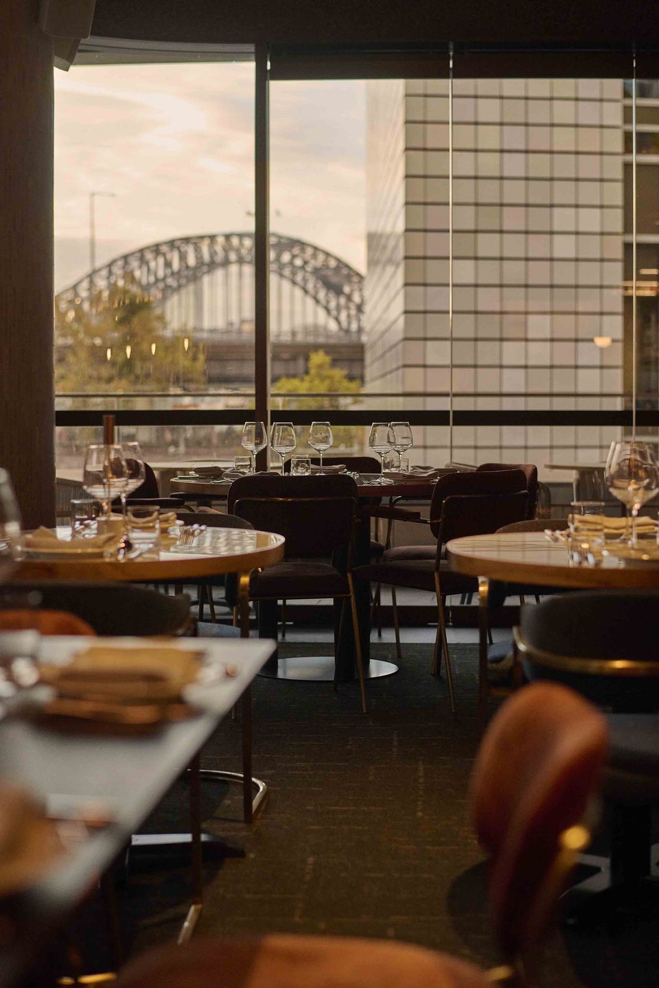 Restaurant interior with tables overlooking a bridge through large windows at sunset