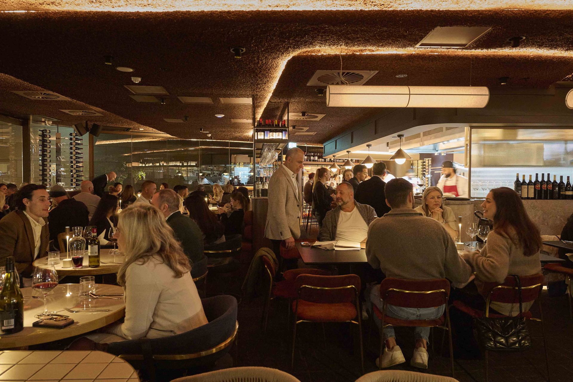 Crowded restaurant with diners seated at tables under warm lighting, chatting and eating.