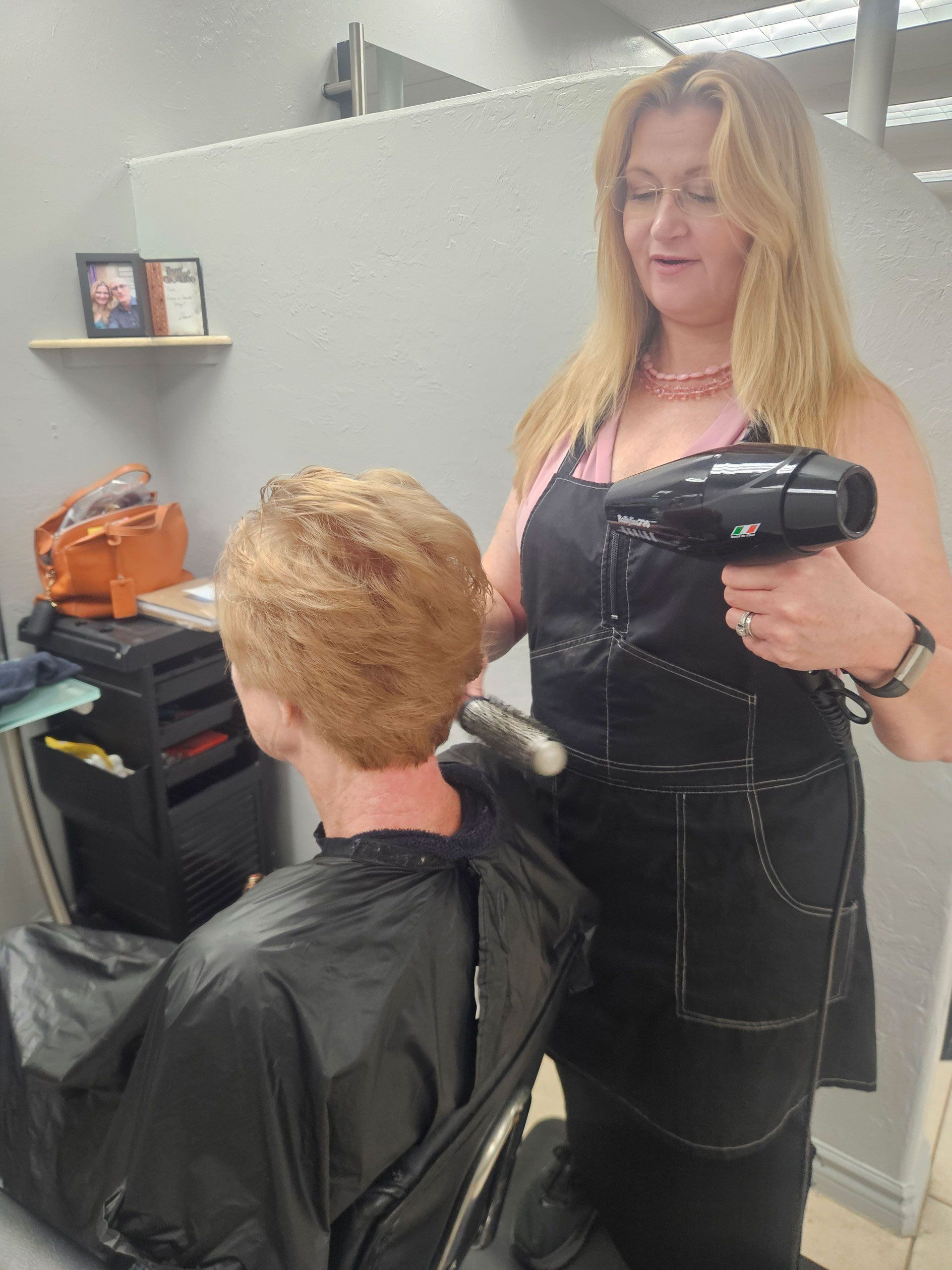Hairdresser blow-drying a client's hair in a salon; the stylist wears a black apron.