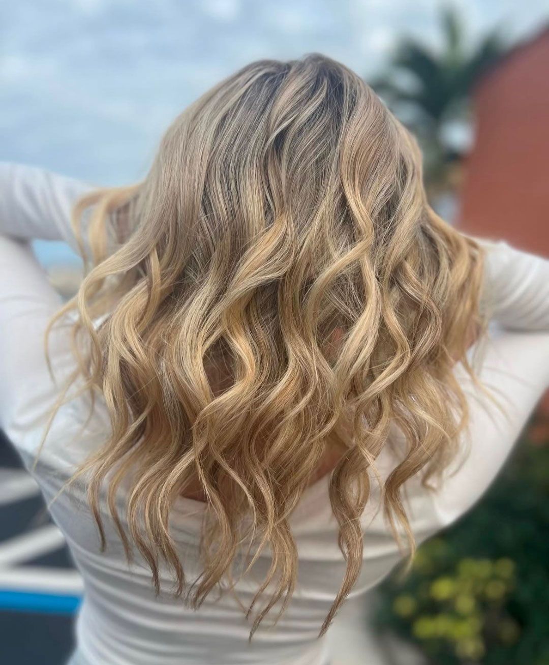 Blonde wavy hair, hands behind head, outdoors.