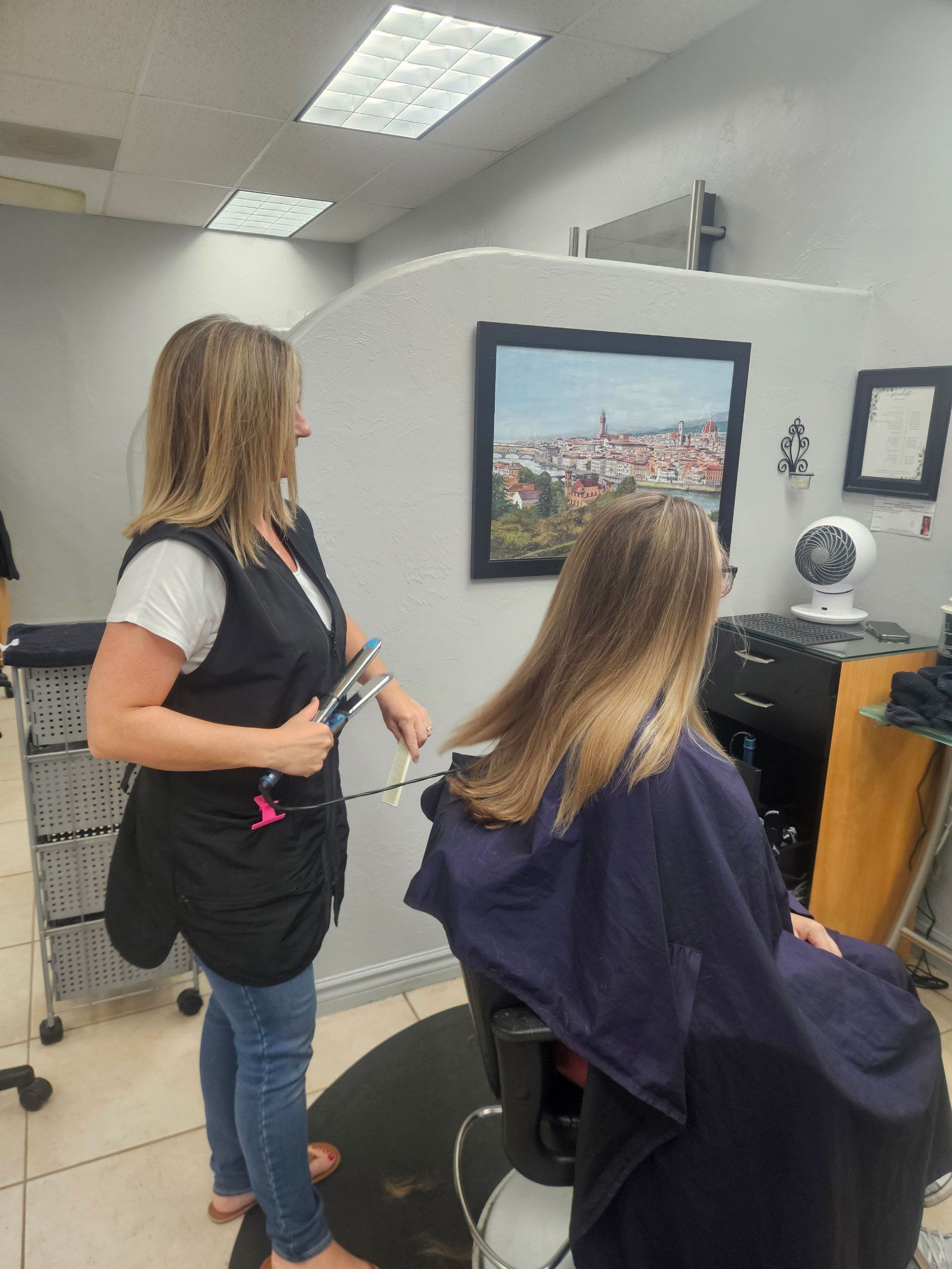 A hairstylist cuts a client's long hair in a salon. The client is seated and draped in a cape.