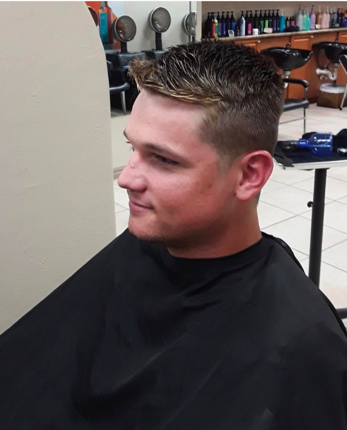 A person in a black salon cape, facing away from the camera in a salon with hair styling equipment visible.