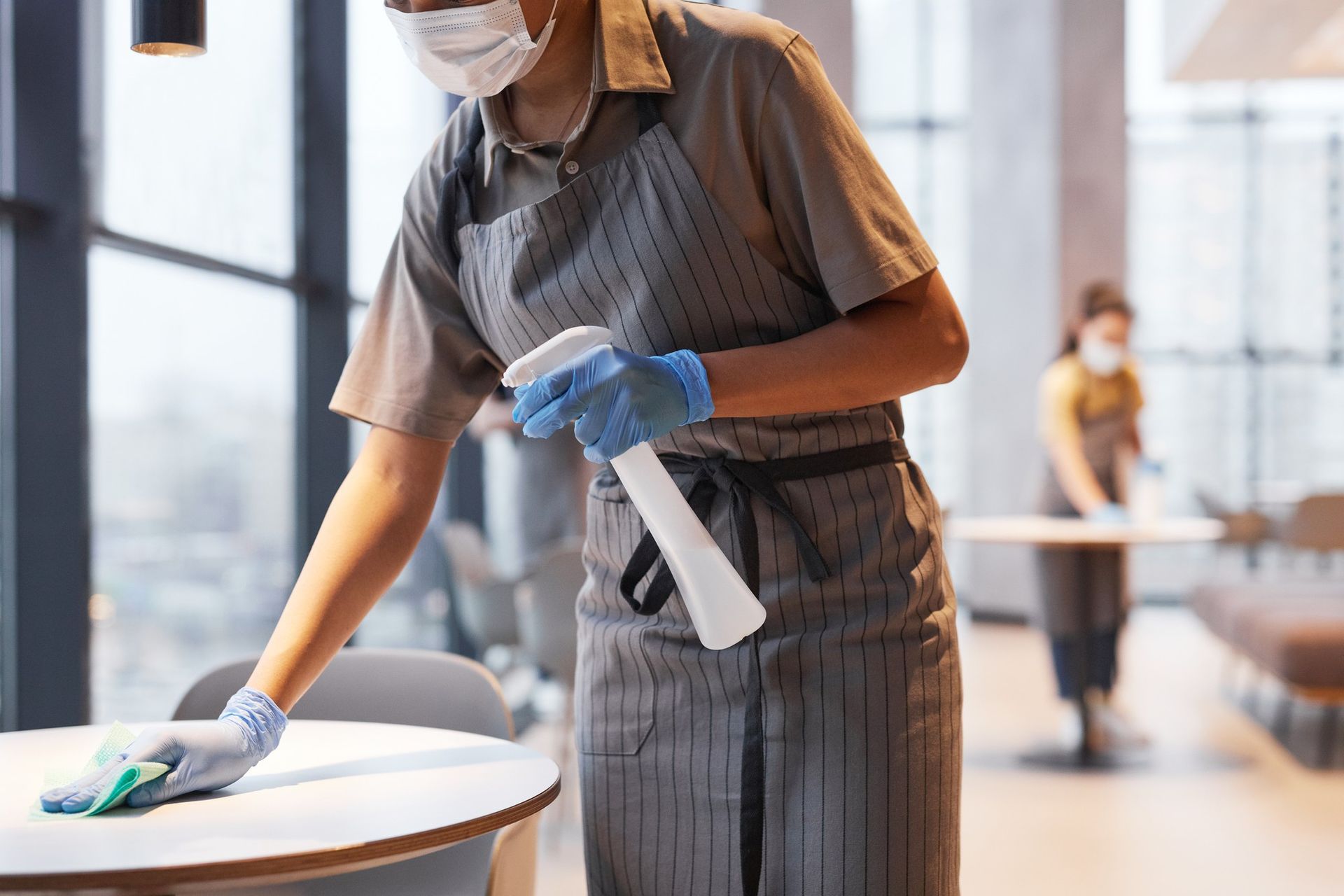 A woman wearing a mask and gloves is cleaning a table in a restaurant.