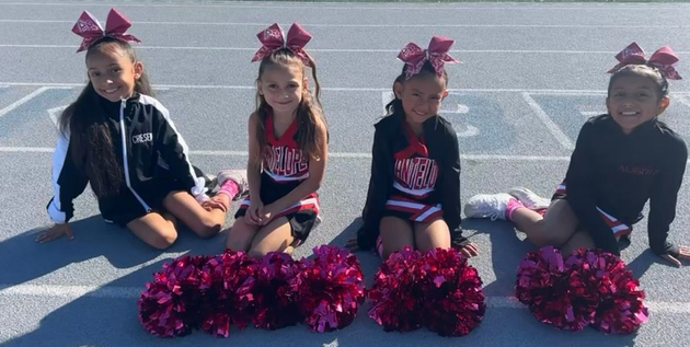 Four cheerleaders sit on pavement with pink pom-poms and bows in front of them.
