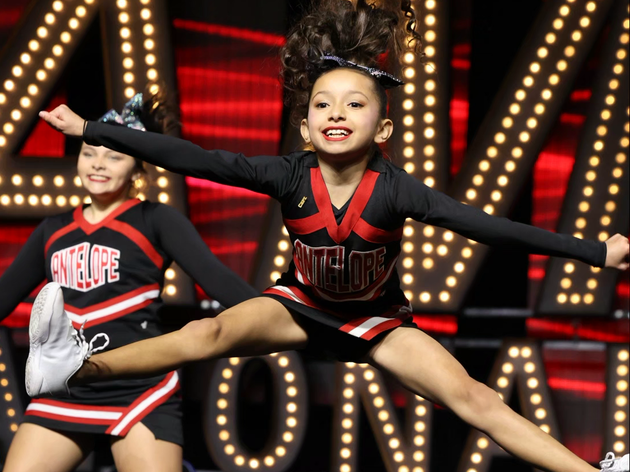 Two cheerleaders in black-and-red uniforms perform high kicks on a lit stage