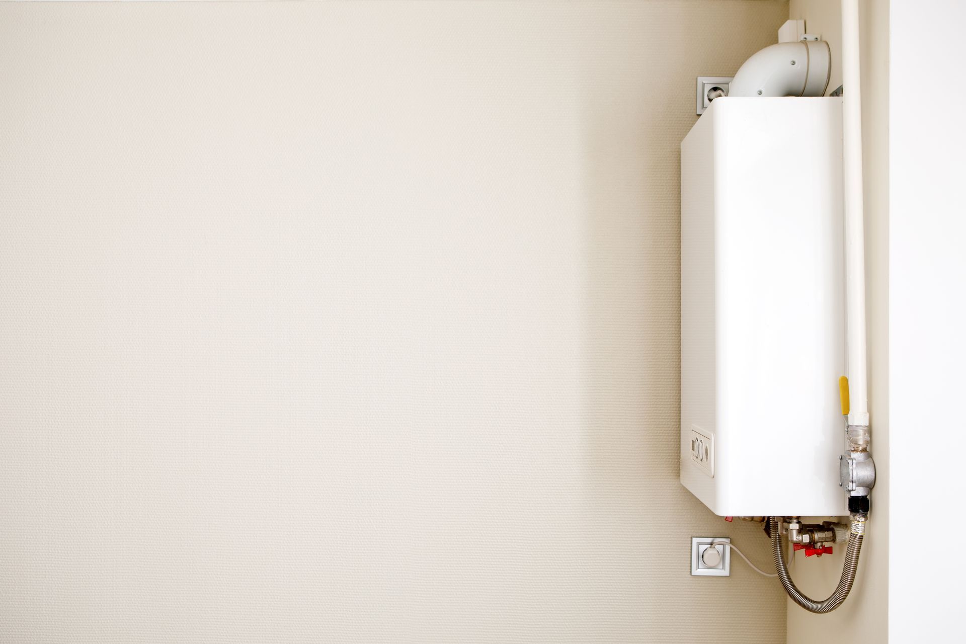 Wall-mounted white water heater on a beige textured wall, with visible pipes and outlet.