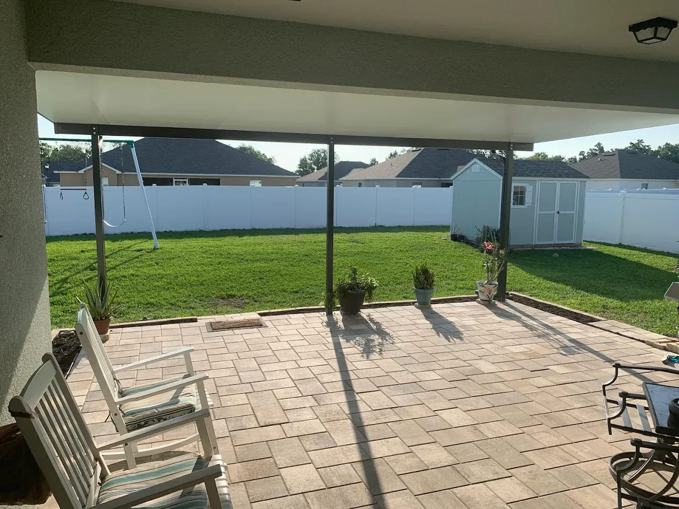 Patio with brick floor, lawn, white fence, shed, and trees. Two chairs on the patio.
