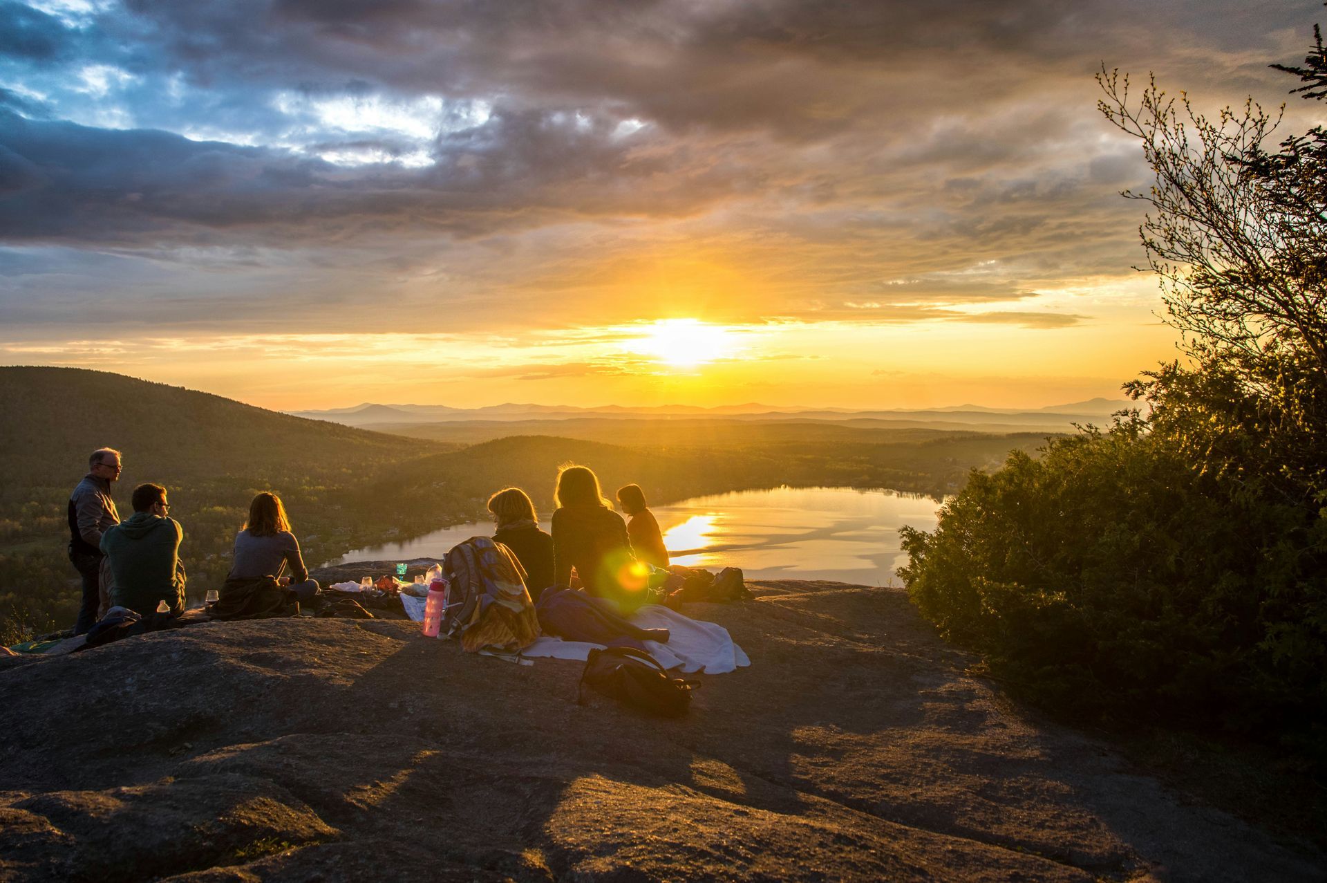 A group of people are sitting on top of a mountain watching the sunset over a lake.