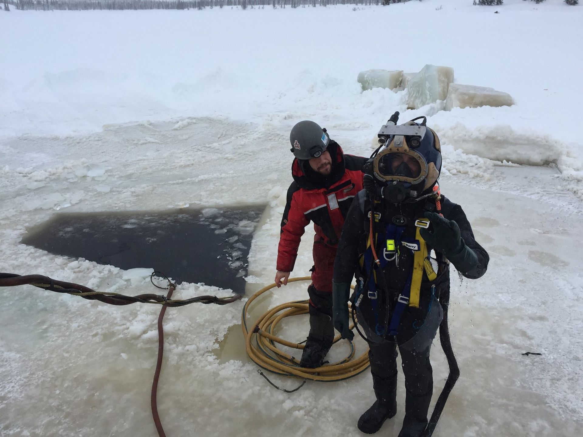 A diver in full gear stands near an ice hole, assisted by a person in a red jacket; snowy landscape.