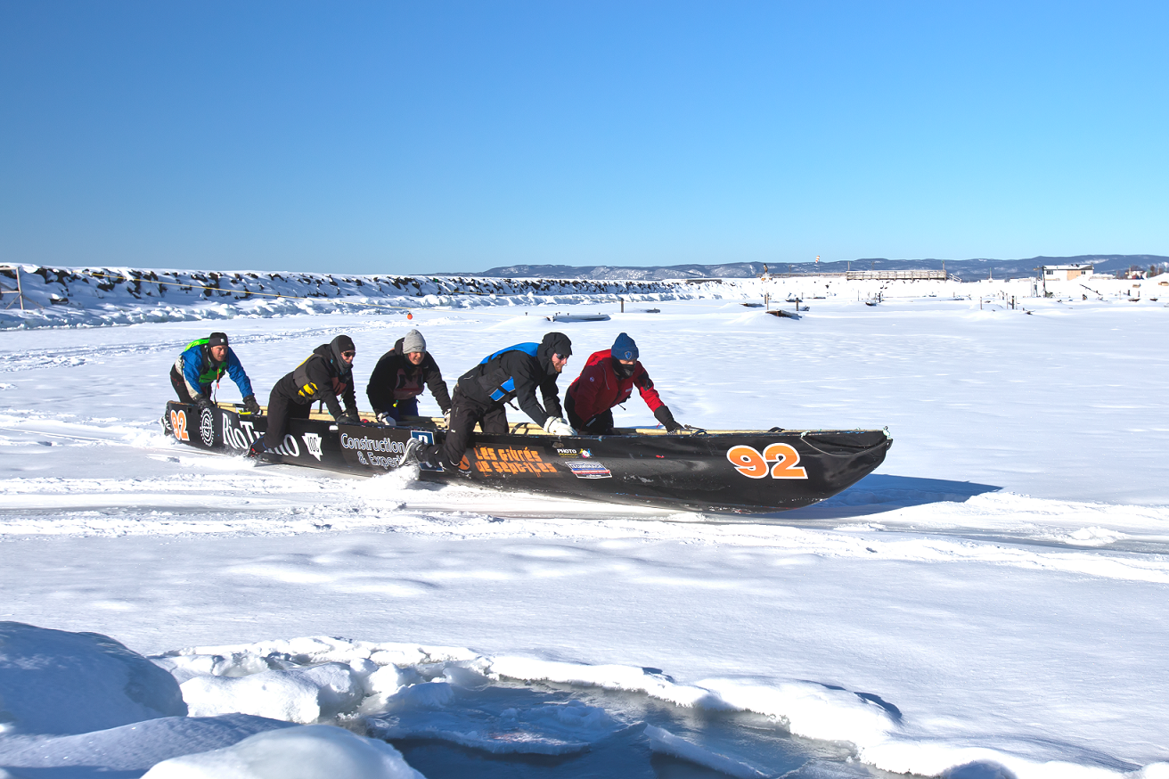 People pushing a black boat across a snowy frozen lake. Blue sky in the background.