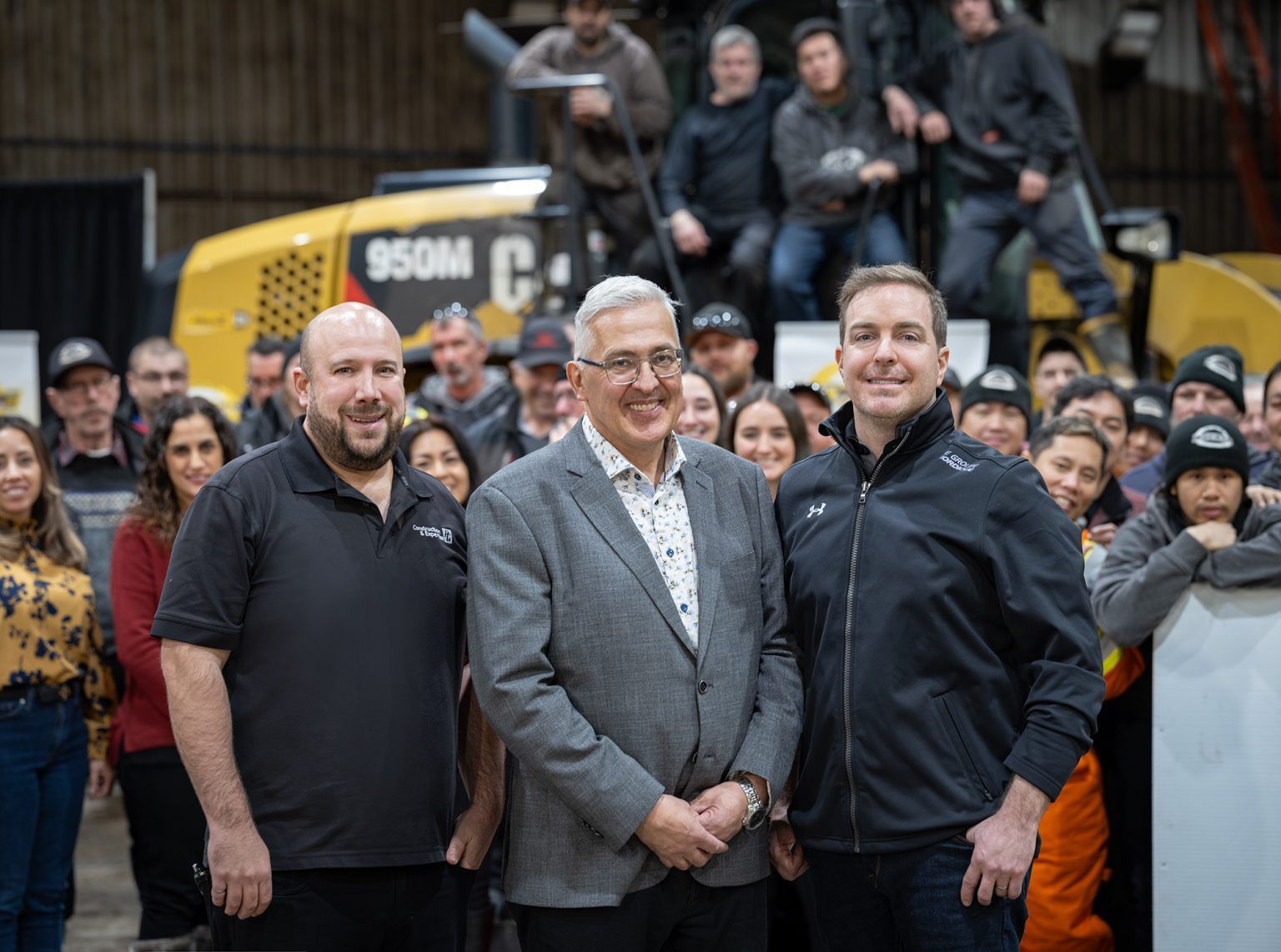 Group of people posing with three men in front, machinery in the background.