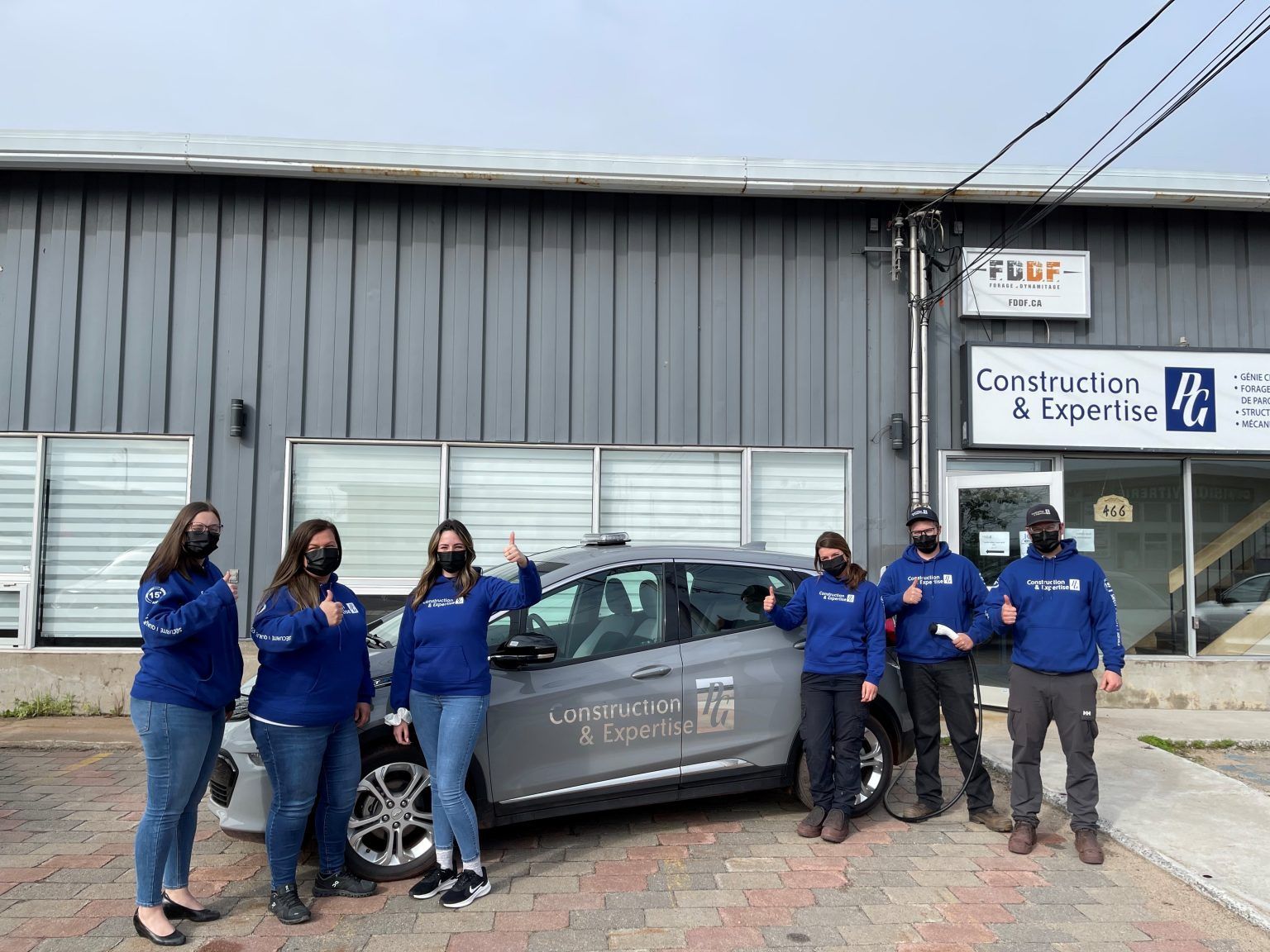 Group of people in matching blue shirts and masks, thumbs up next to a car with company logo in front of a building.