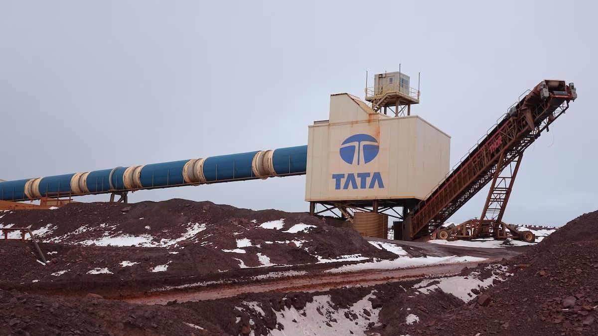 Industrial facility with a conveyor belt and pipe, branded with the TATA logo, over a pile of reddish-brown material, outdoors.