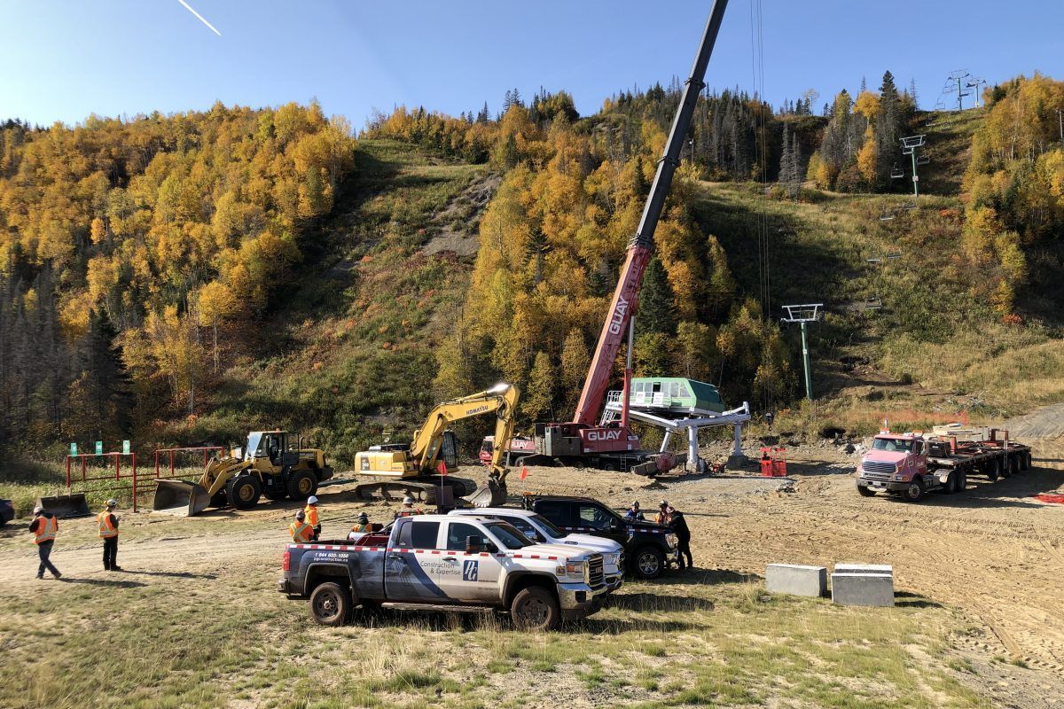 Construction site on a mountain slope with machinery, crane, trucks, and workers. Autumn foliage in background.