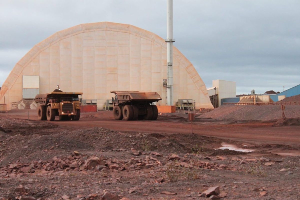 Two large dump trucks driving on a red dirt road, with a large white arched building in the background.