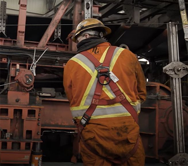 Un ouvrier en combinaison orange et casque de chantier, portant un harnais de sécurité, fait face à une machine à l'intérieur d'une usine.