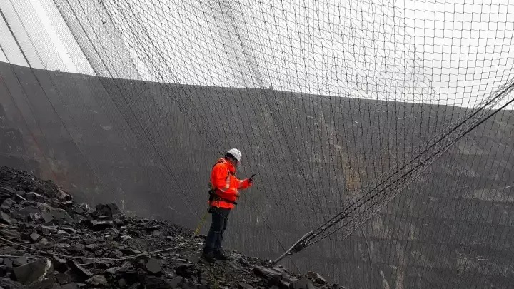 Man in orange jacket inspects large protective net in a quarry.